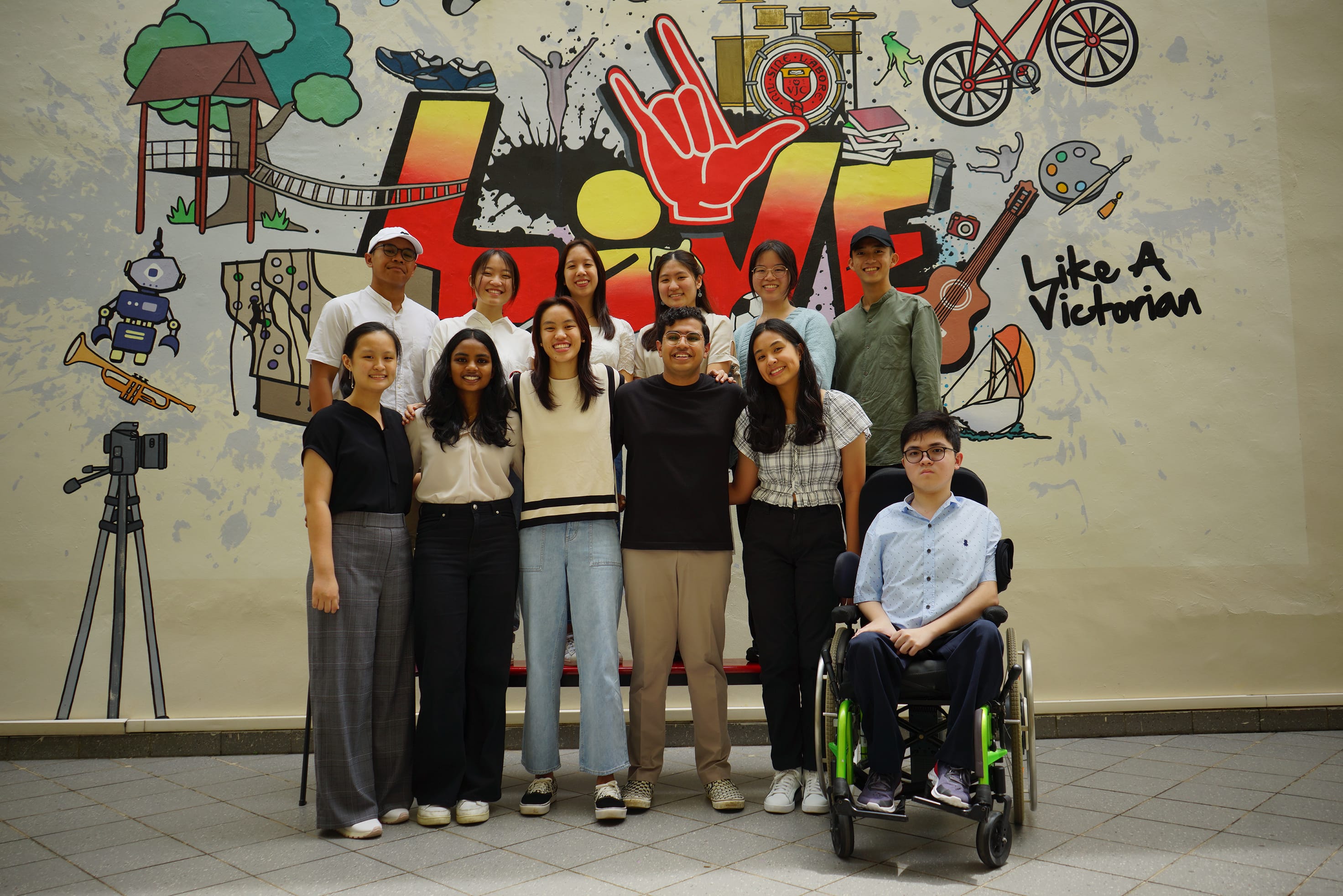 A group of students and staff posing for a photo in front of a colorful mural that prominently features the word “LOVE” and the phrase “Like a Victorian,” along with illustrated icons such as a bicycle, books, a guitar, and a camera. The group is arranged in two rows, smiling at the camera; one person is seated in a wheelchair at the front right.